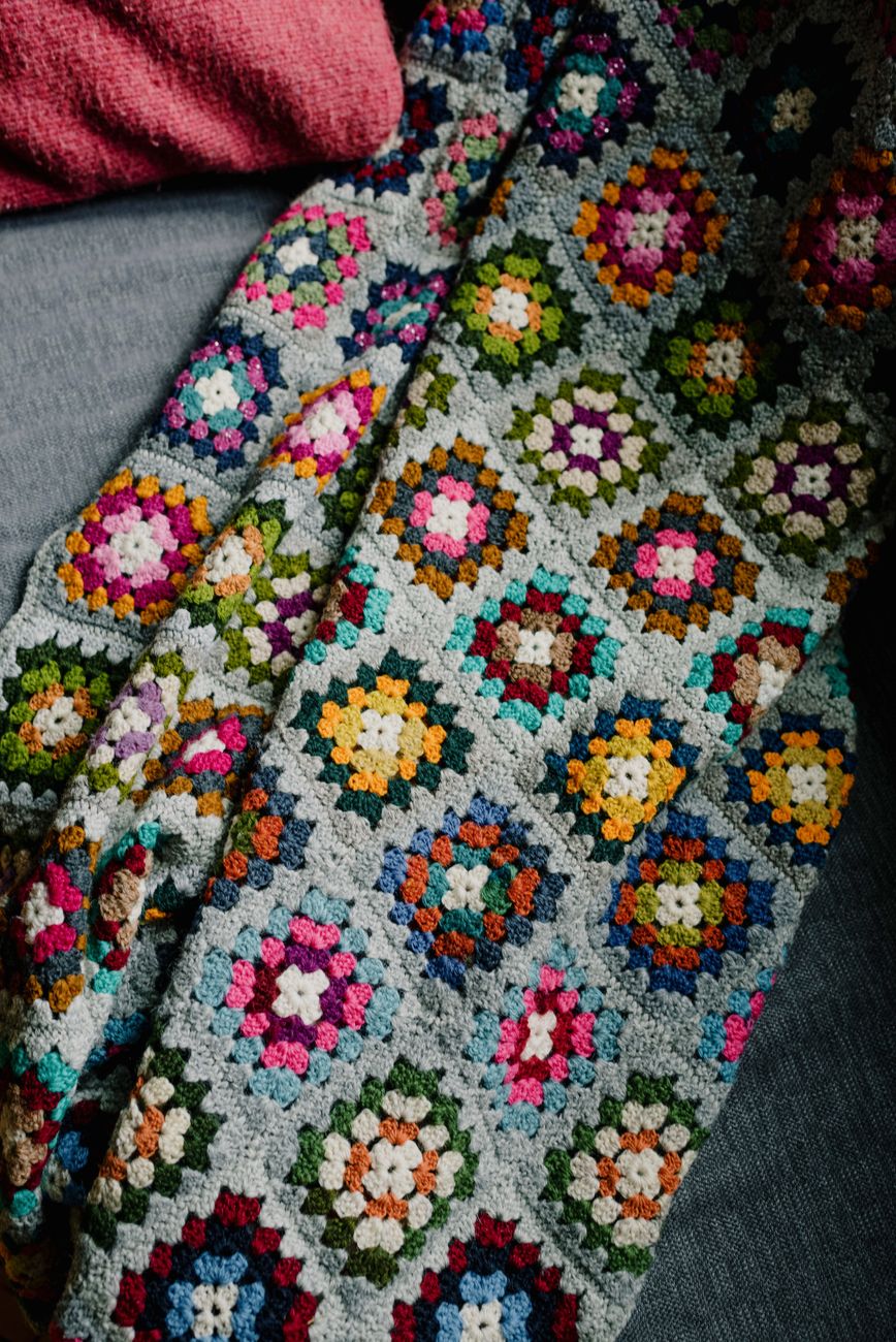 Close-up of a colorful crocheted blanket featuring a geometric pattern with floral motifs in various colors, laid over a gray surface.